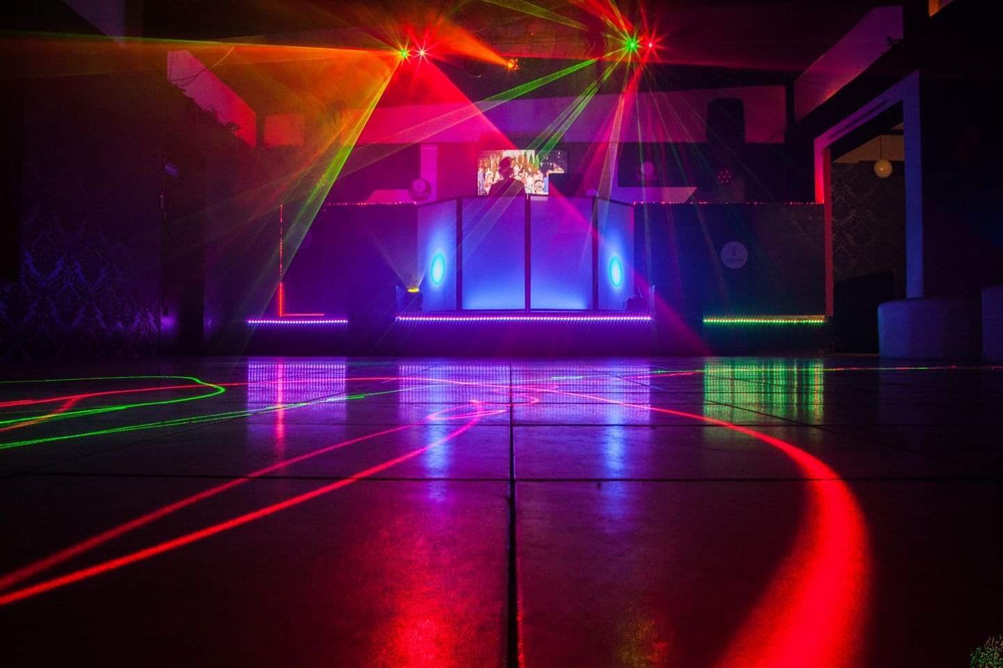 Parkiet oświetlony laserami w kolorach czerwonym i zielonym, w tle stanowisko DJ-a z ekranem i dodatkowym oświetleniem w kolorach niebieskim i fioletowym.