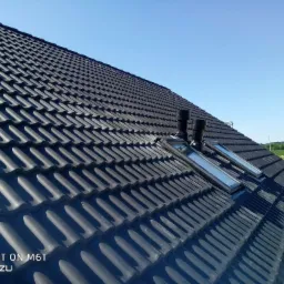 Nowy dach z czarnej dachówki ceramicznej z zamontowanymi oknami dachowymi i kominkami wentylacyjnymi, widok z dołu w słoneczny dzień.