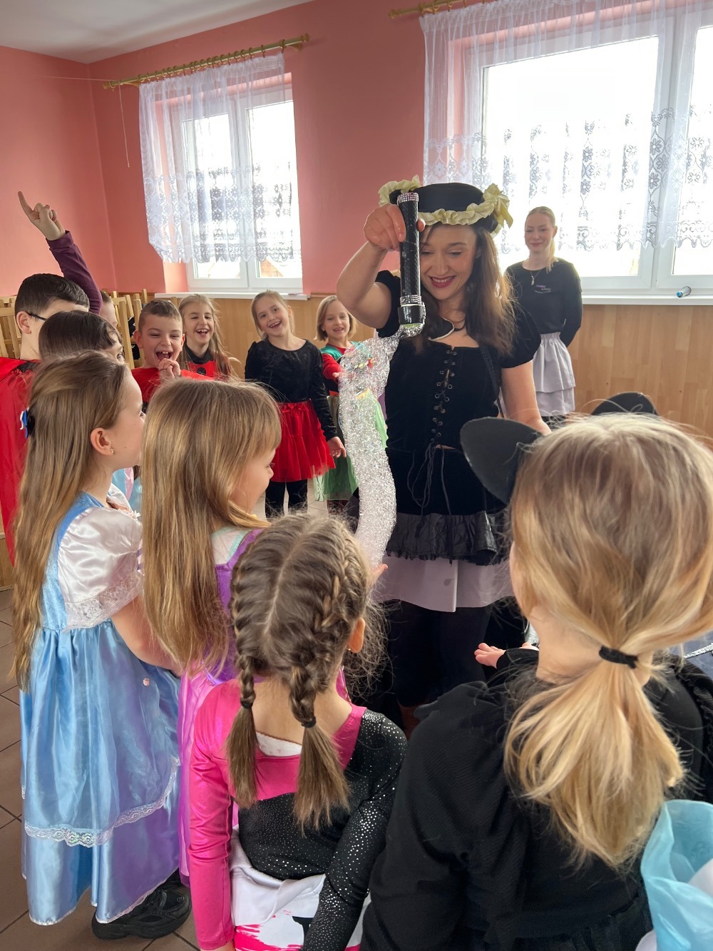 Kobieta w kostiumie pirata prezentuje dzieciom urządzenie do robienia baniek mydlanych podczas imprezy w sali z oknami.