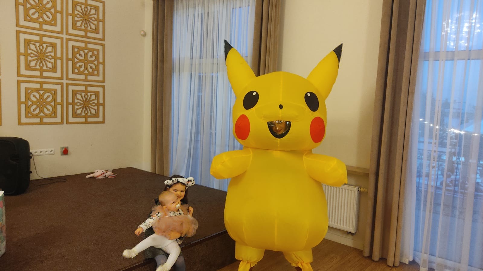 Żółty, dmuchany kostium Pikachu stoi na scenie, obok siedząca dziewczynka trzymająca niemowlę w różowej tiulowej spódniczce.