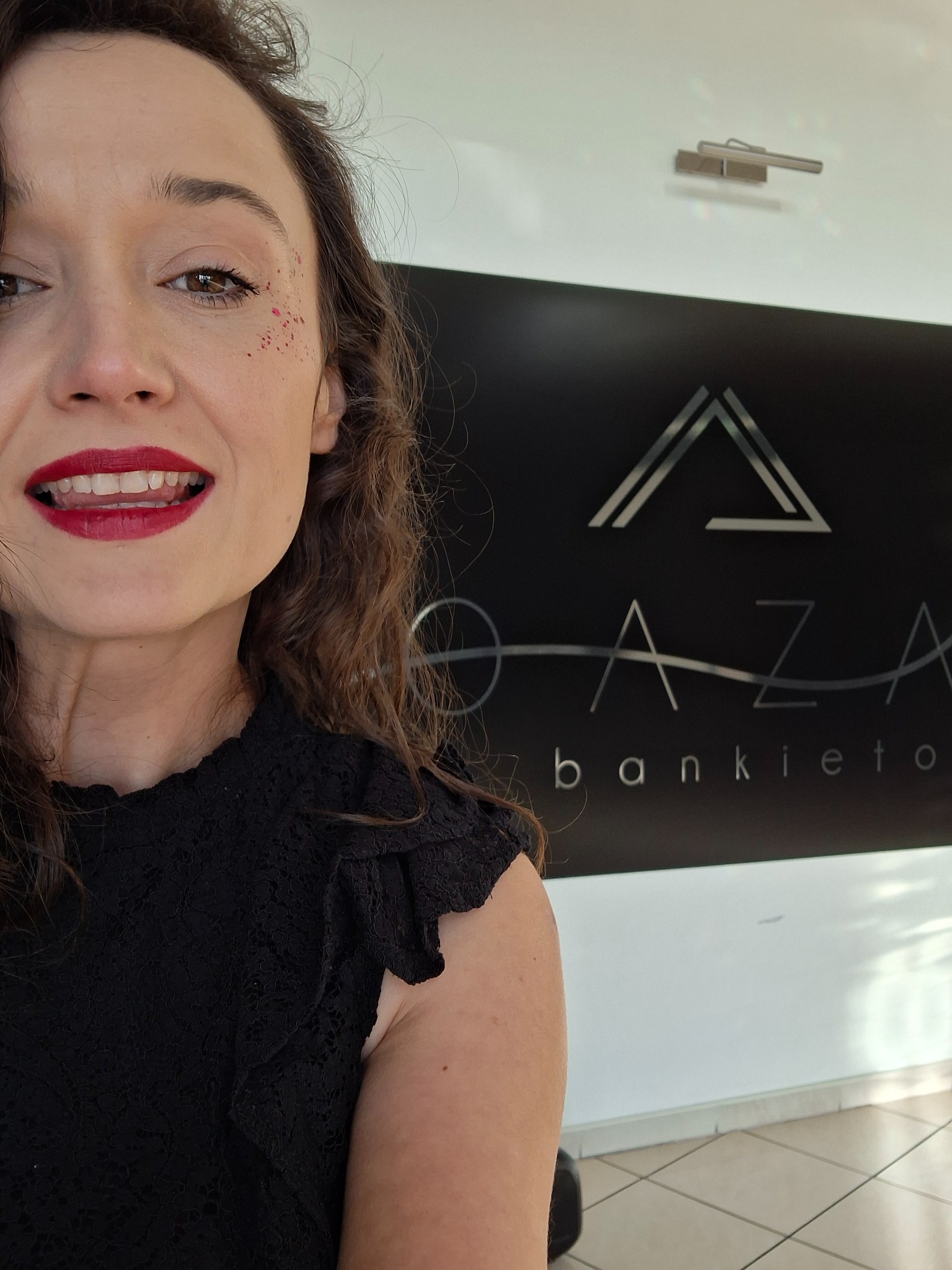 Uśmiechnięta kobieta z czerwonymi ustami i brokatem na policzku, na tle logo 'OAZA bankietowa'.