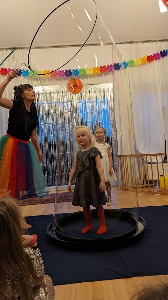 Dziewczynka w sukience i czerwonych rajstopach stoi wewnątrz gigantycznej bańki mydlanej tworzonej przez kobietę w tęczowej spódnicy, w tle dekoracje imprezowe.