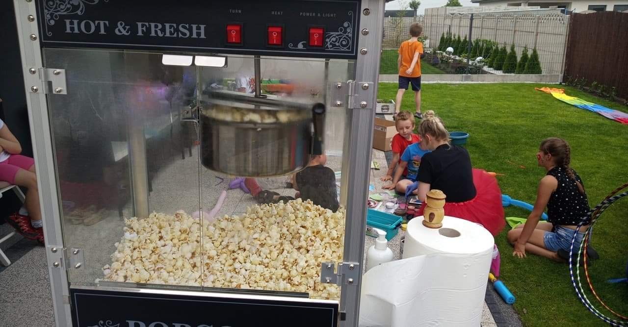 Automat do popcornu 'Hot & Fresh' na trawniku podczas imprezy dla dzieci, w tle bawiące się dzieci i ogrodzenie.