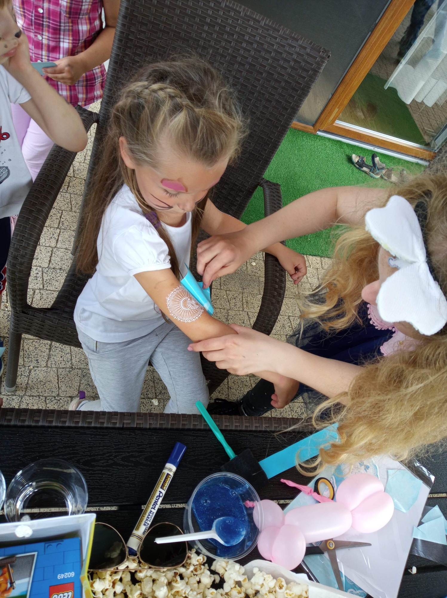 Dziewczynka z pomalowaną twarzą i wzorem henny na ramieniu, wykonywanym przez osobę z opaską z motylami, na tle stolika z popcornem, slime i balonami.