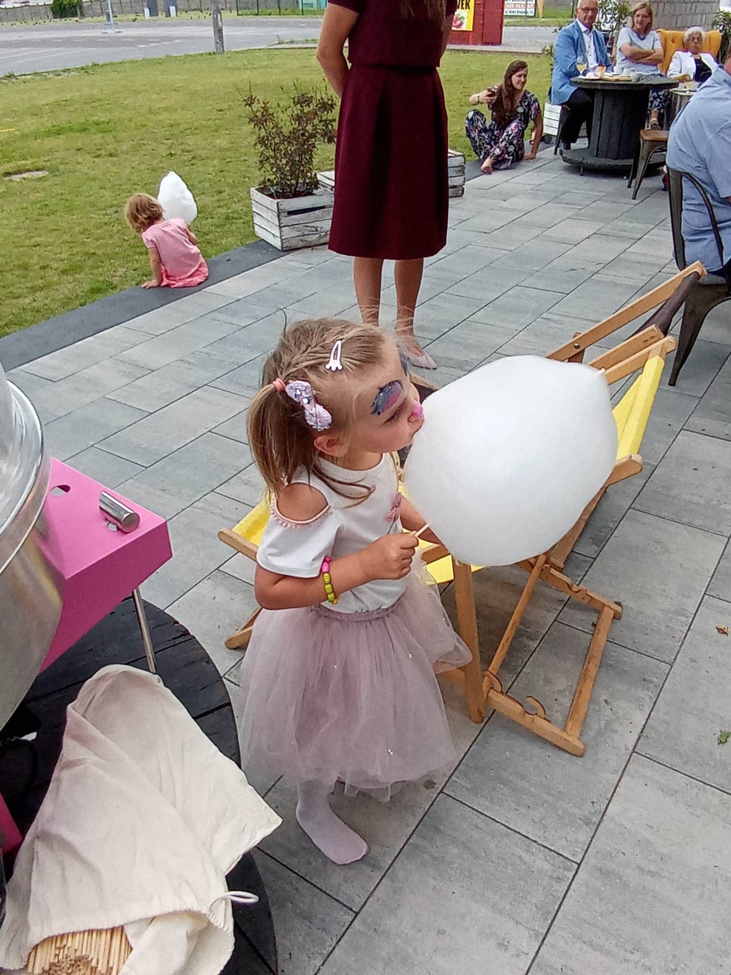 Dziewczynka z pomalowaną twarzą, w tiulowej spódniczce, siedzi na małym krzesełku i je watę cukrową podczas imprezy plenerowej w Lesznie; w tle widoczni goście.