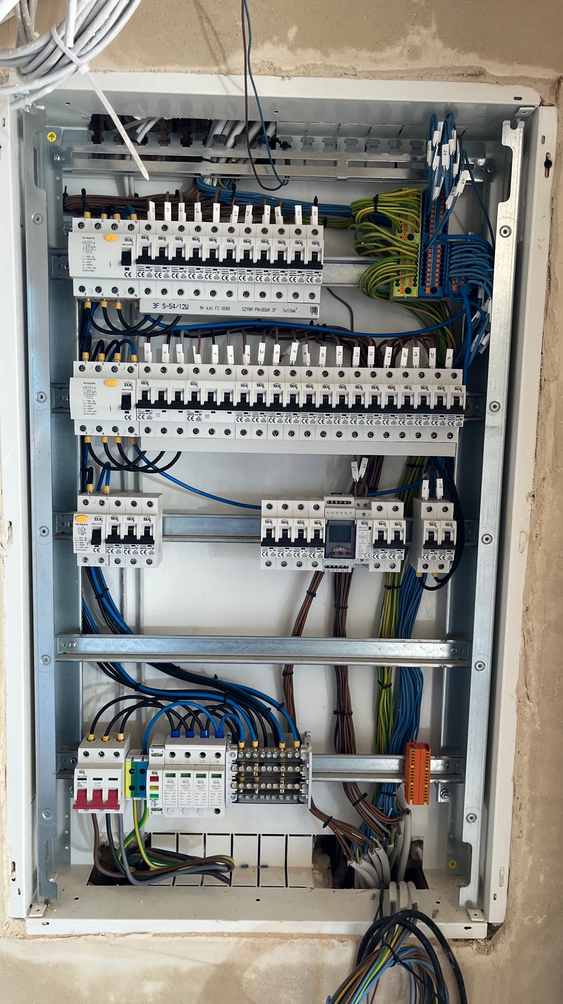 Rozdzielnica elektryczna w metalowej obudowie, widoczne zabezpieczenia, przewody i listwy zaciskowe. Instalacja w trakcie montażu, surowy stan wykończenia ściany.