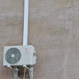 Zamontowana jednostka zewnętrzna klimatyzacji marki Rotenso z białymi rurami odprowadzającymi skropliny, przymocowana do szarej, chropowatej ściany budynku.