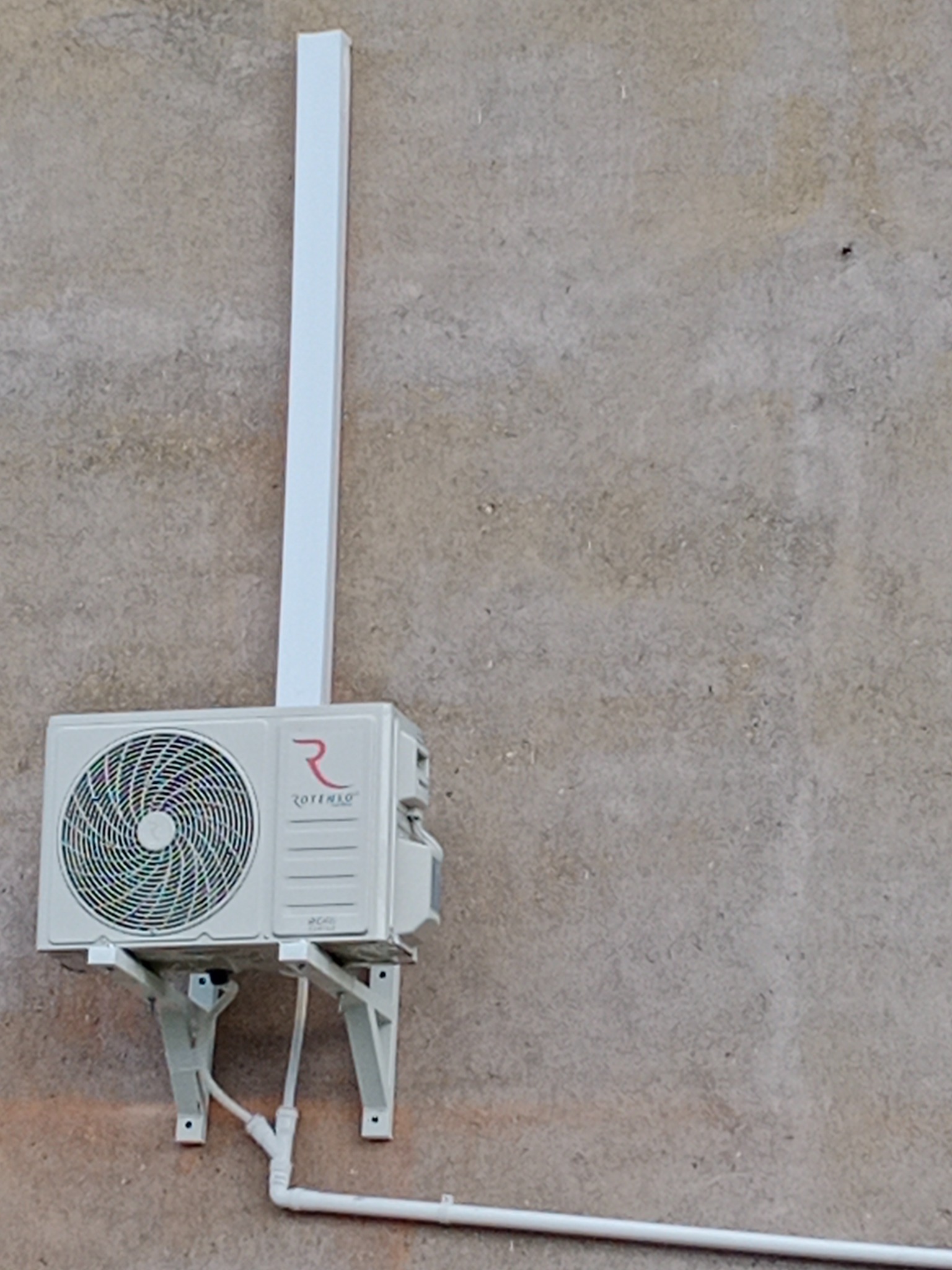 Zamontowana jednostka zewnętrzna klimatyzacji marki Rotenso z białymi rurami odprowadzającymi skropliny, przymocowana do szarej, chropowatej ściany budynku.