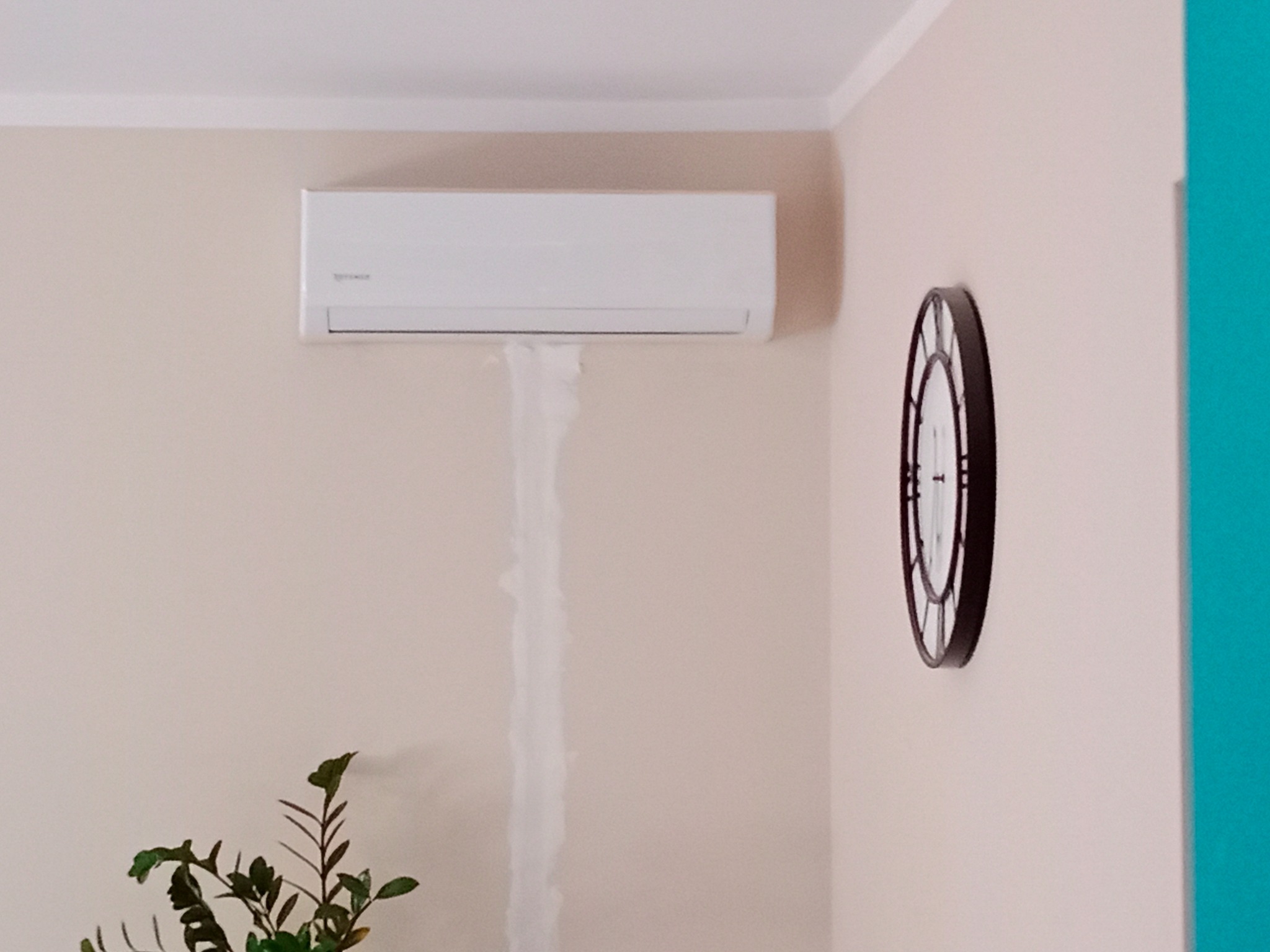 Biały klimatyzator ścienny w salonie z zegarem i rośliną doniczkową, widoczne ślady wyciekającej wody.