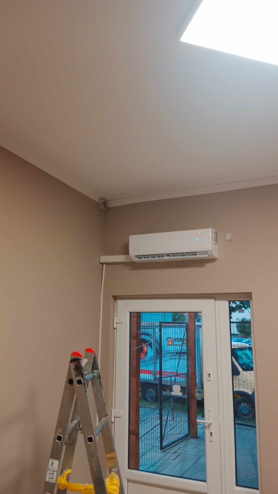 Biała jednostka klimatyzacyjna zamontowana nad drzwiami, widoczna drabina aluminiowa z żółtymi elementami zabezpieczającymi, beżowe ściany i biały sufit.