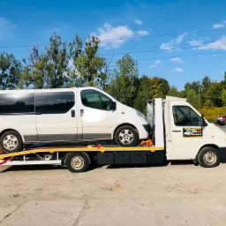 Biały bus na platformie lawety na tle zieleni i błękitnego nieba. Na drzwiach lawety naklejka z napisem 'Pomoc Drogowa'.