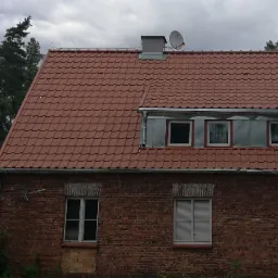 Ceglany dom z anteną satelitarną na dachu z czerwonej dach&oacute;wki, widoczną na tle pochmurnego nieba i drzew.