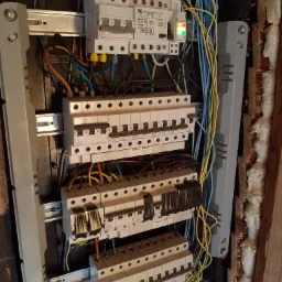 Rozdzielnia elektryczna z widocznymi bezpiecznikami, przewodami w różnych kolorach (niebieski, żółty, pomarańczowy, czarny) oraz elementami montażowymi, zamontowana w ścianie z widoczną pianką...