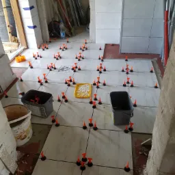 Układanie jasnych płytek podłogowych z użyciem systemu poziomowania płytek, widoczne dwa wiadra i kratka ściekowa