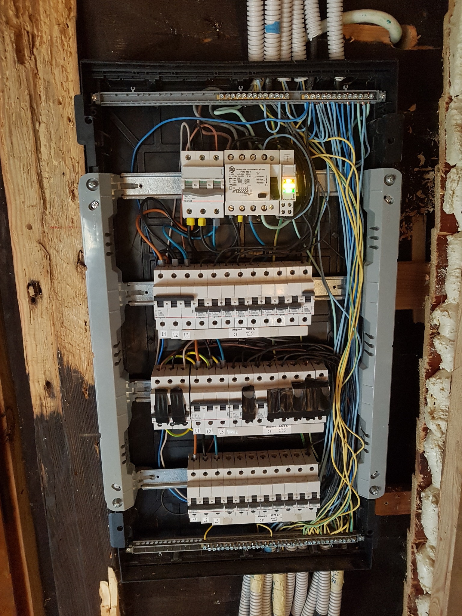 Rozdzielnica elektryczna z zainstalowanymi bezpiecznikami i przewodami w trakcie montażu, widoczne kable w kolorach niebieskim i żółtym, oraz fragment drewnianej ściany z pianką montażową.