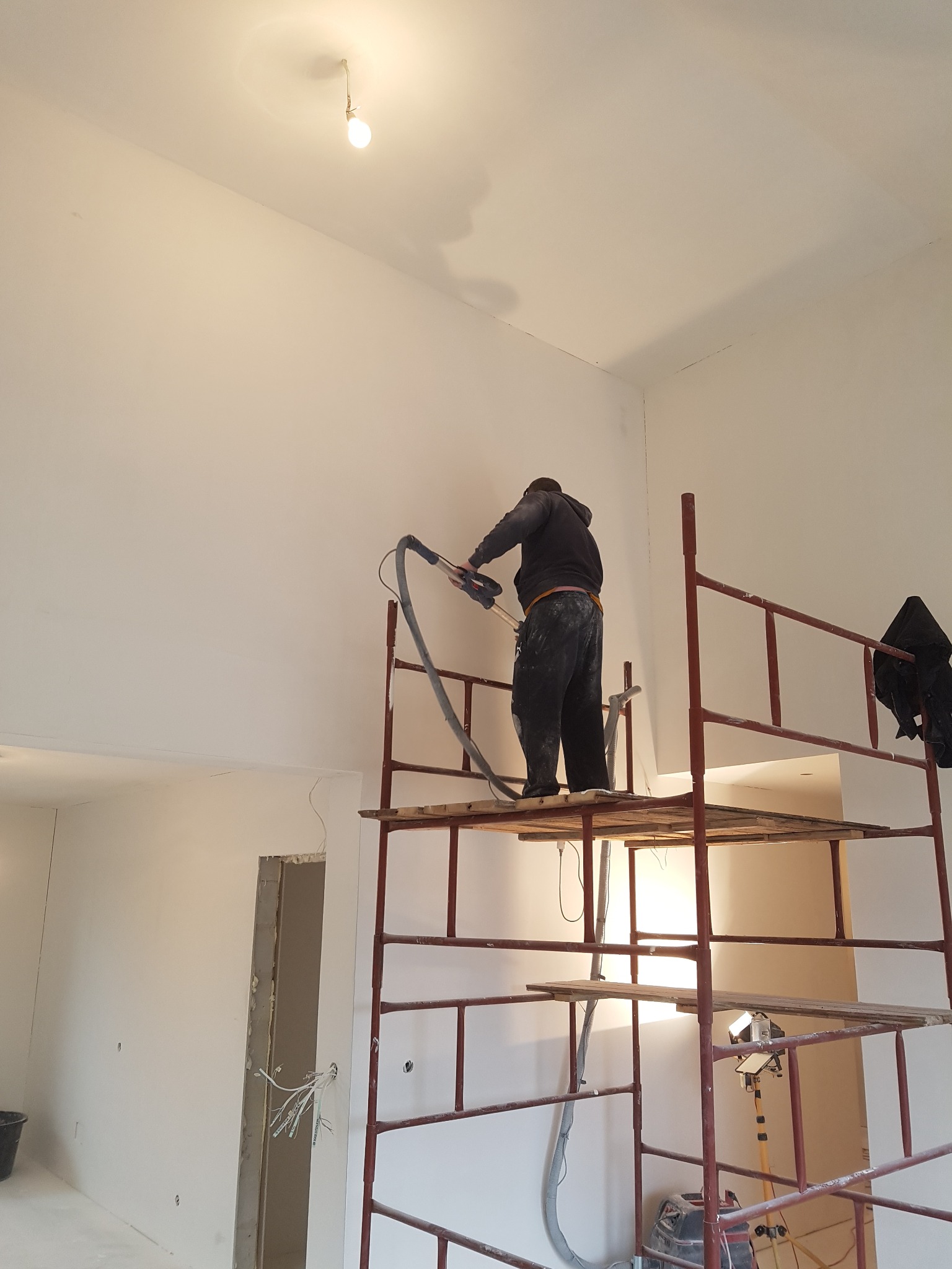 Pracownik na rusztowaniu szlifuje ścianę w wysokim pomieszczeniu, używając szlifierki z odkurzaczem, widoczne przewody elektryczne w ścianie