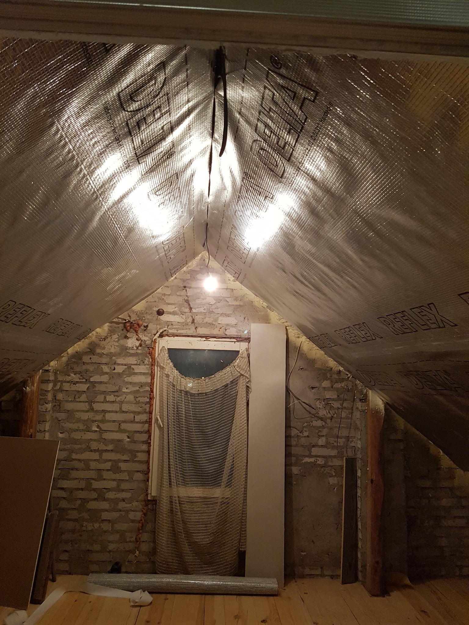 Poddasze w trakcie prac, widoczna folia termoizolacyjna z nadrukiem, surowa cegła, okno z firanką i drewniana podłoga, przygotowane materiały budowlane.