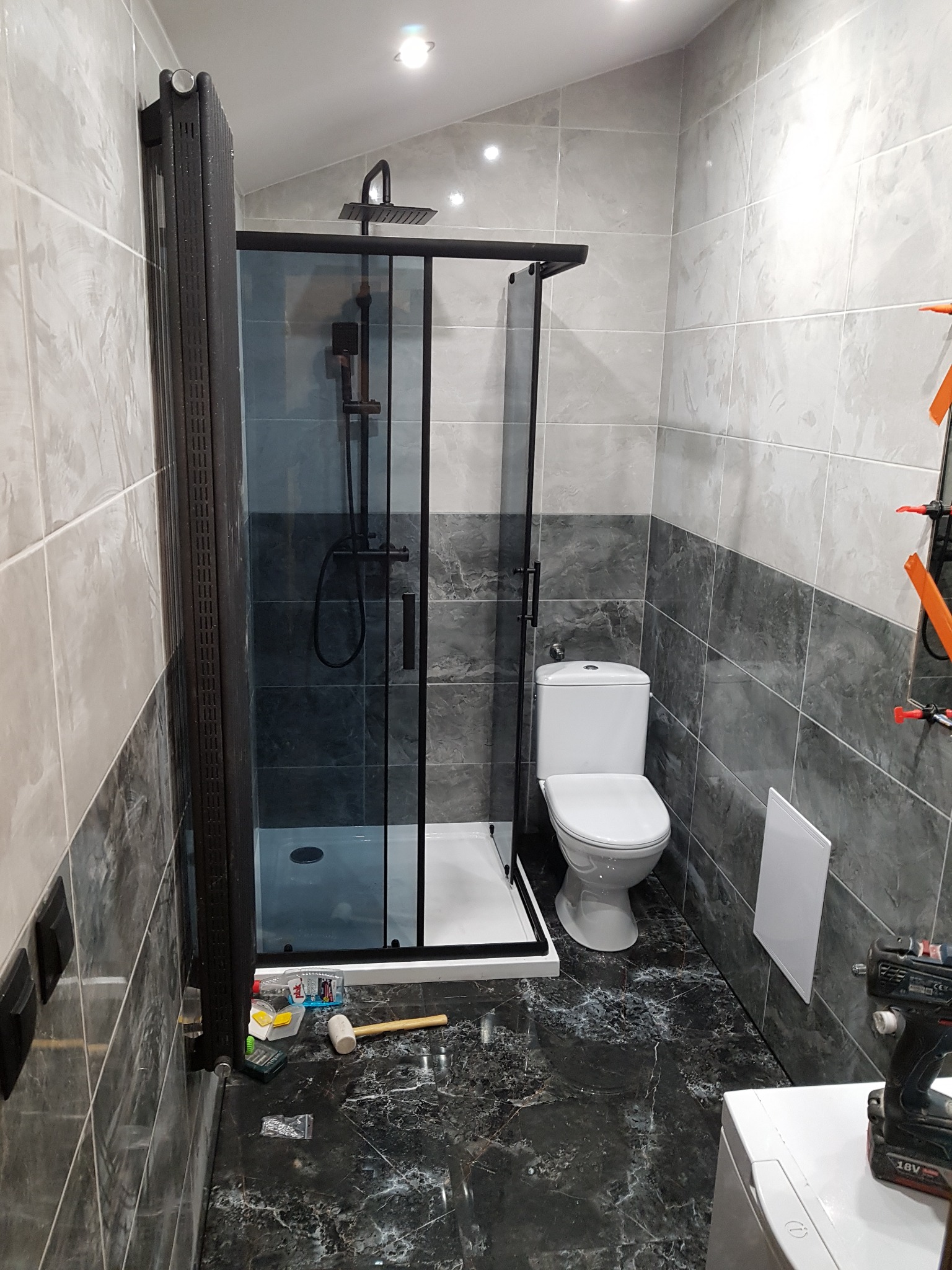 Wykończona łazienka z czarną kabiną prysznicową typu walk-in, białą toaletą i marmurową podłogą, widoczne narzędzia i akcesoria glazurnicze.