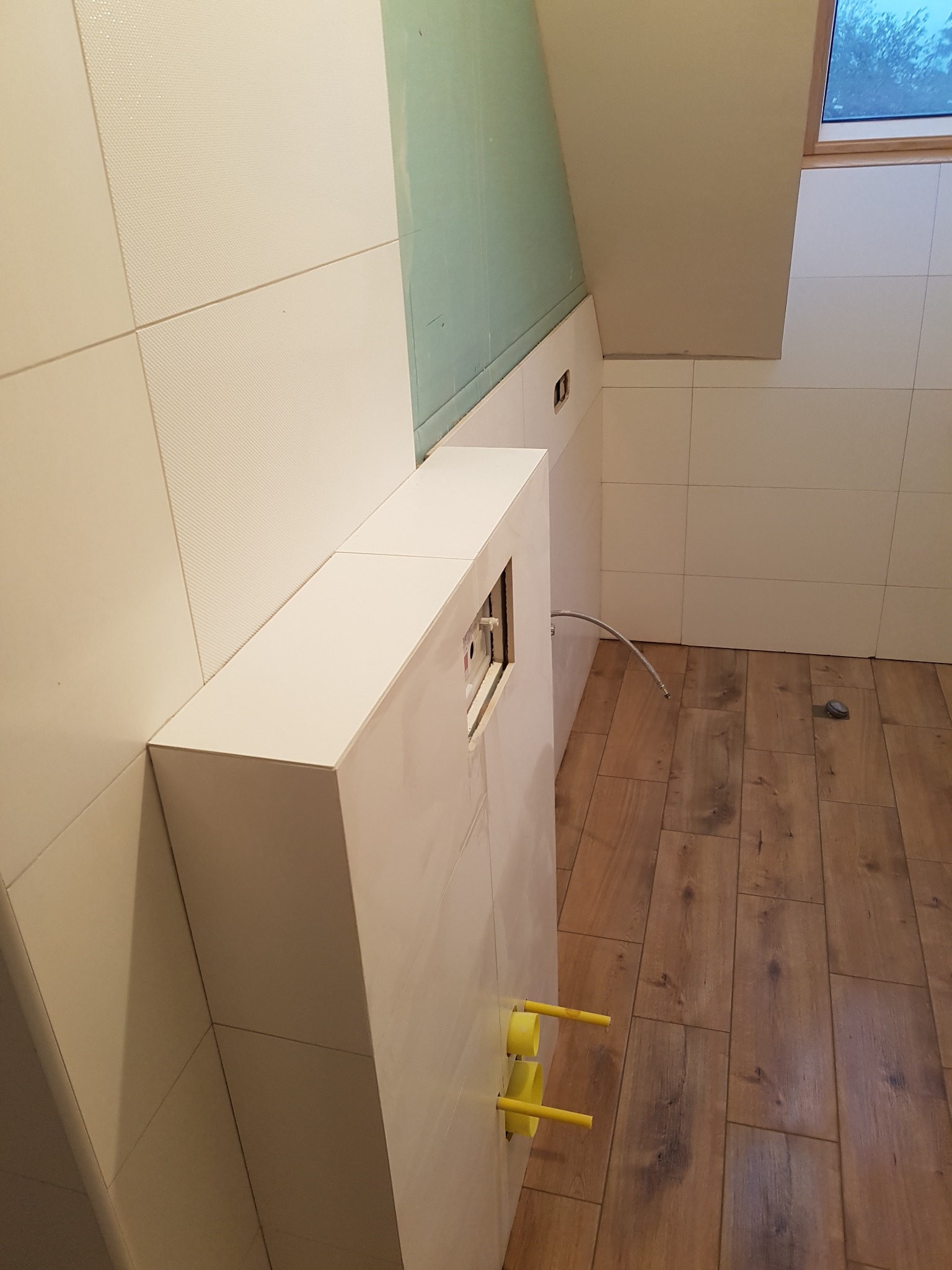 Białe płytki ścienne z delikatną fakturą w łazience na poddaszu, zabudowa stelaża podtynkowego WC z widocznymi żółtymi rurami, drewnopodobne panele podłogowe.