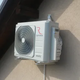 Zewnętrzna jednostka klimatyzacji Rotenso Inverter zamontowana na ścianie budynku, widoczna instalacja elektryczna i rura odprowadzająca skropliny, tło stanowi fragment dachu i antena satelitarna.