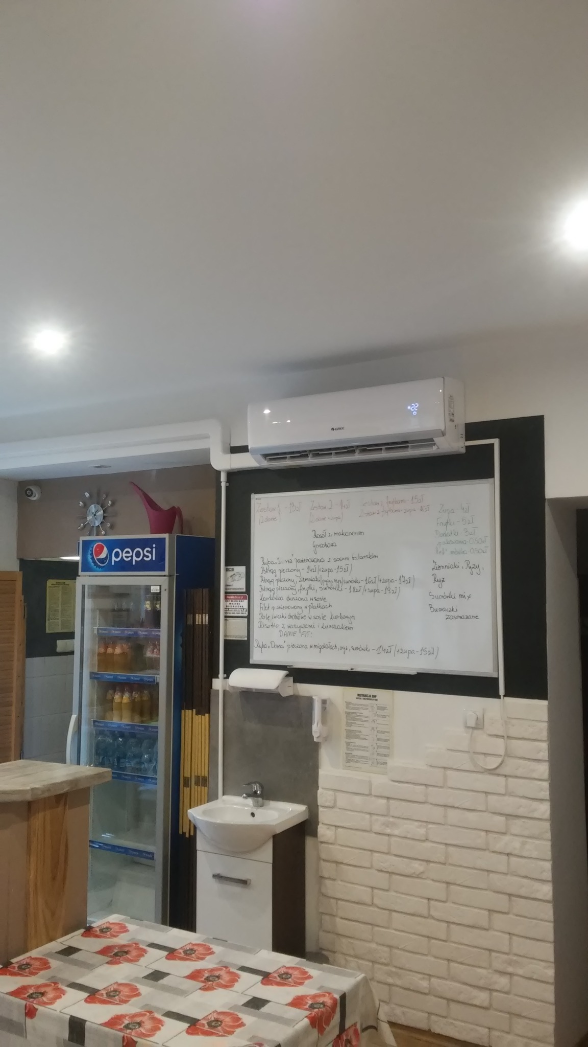 Wnętrze lokalu gastronomicznego z zainstalowanym klimatyzatorem marki Gree nad tablicą z menu, obok umywalki i lodówki Pepsi.