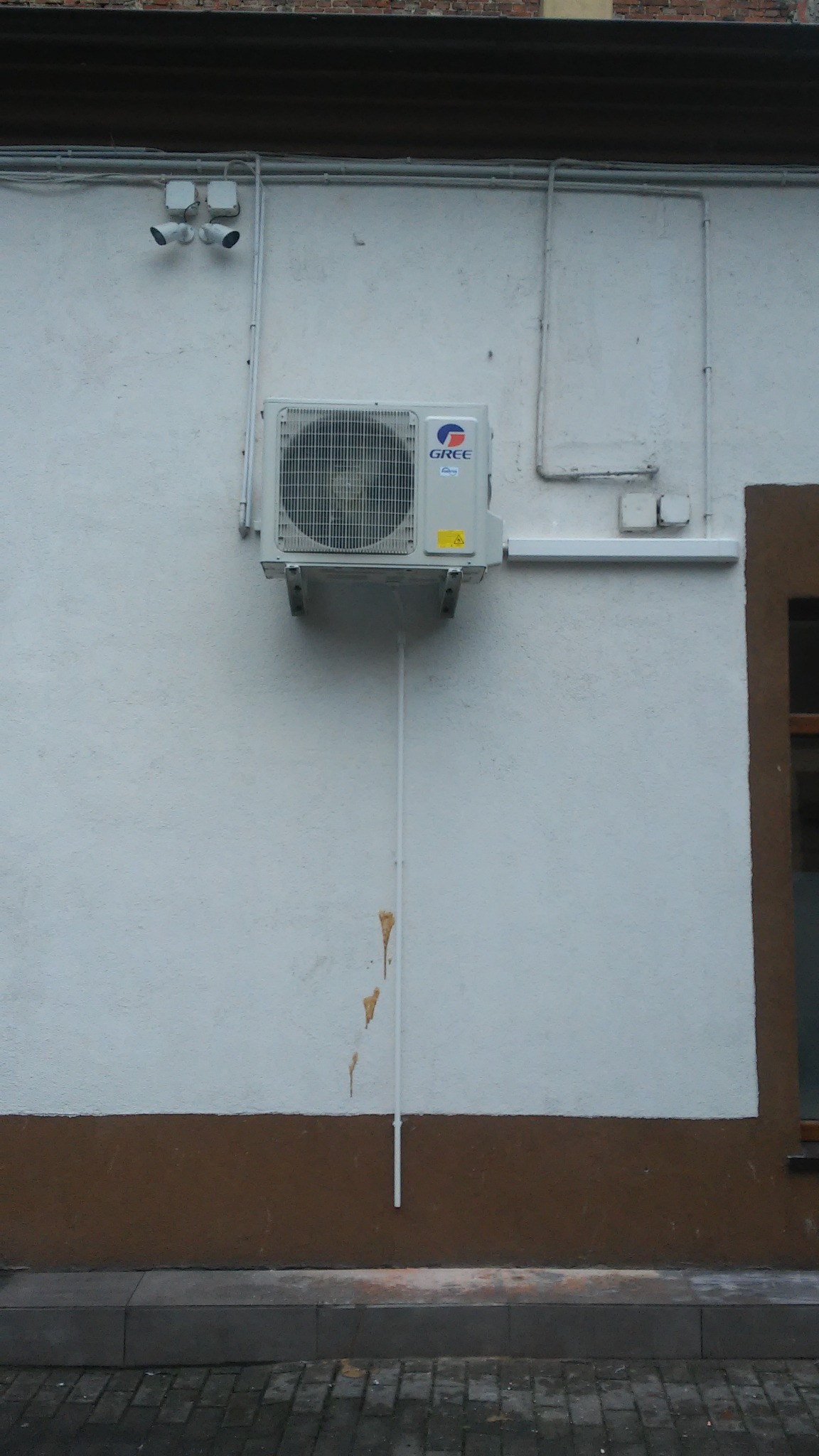 Zewnętrzna jednostka klimatyzacji marki Gree zamontowana na białej, szorstkiej ścianie budynku, widoczne rury instalacyjne i kamery monitoringu.