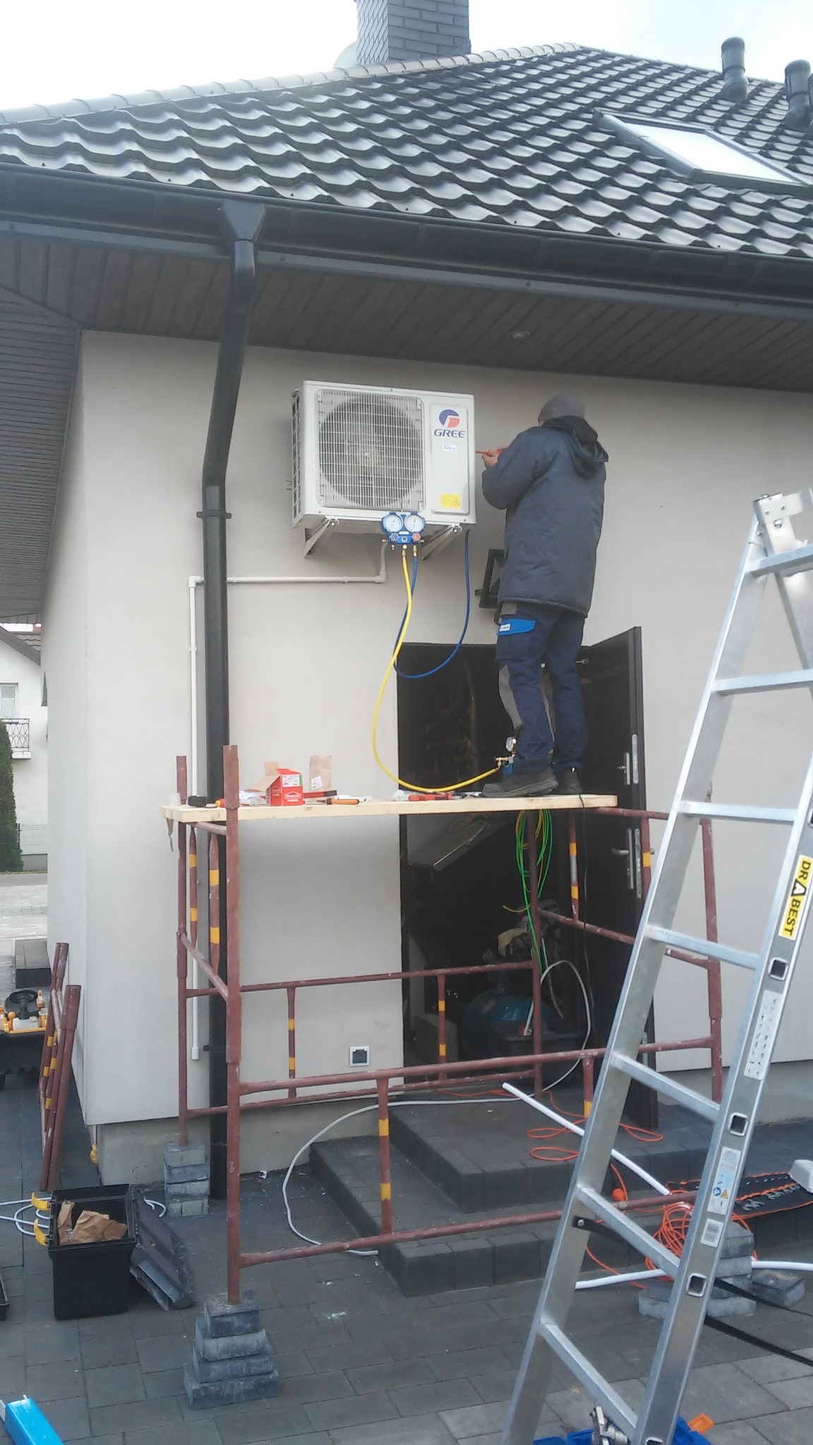Pracownik w kurtce i czapce, na rusztowaniu, podłącza manometry do zewnętrznej jednostki klimatyzatora marki Gree zamontowanej na ścianie domu z szarą dachówką.