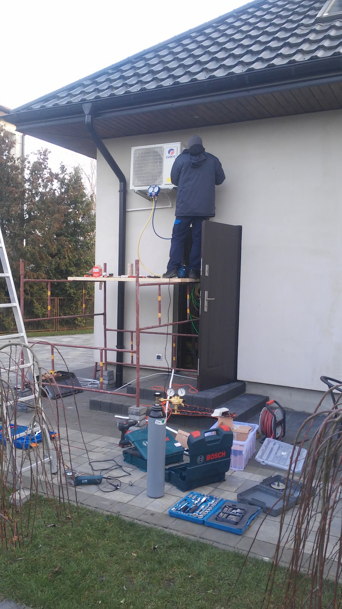 Mężczyzna w kurtce roboczej stoi na rusztowaniu przed budynkiem, podłączając manometry do zewnętrznej jednostki klimatyzatora na ścianie. Na ziemi widoczne narzędzia i butla gazowa, sugerujące...