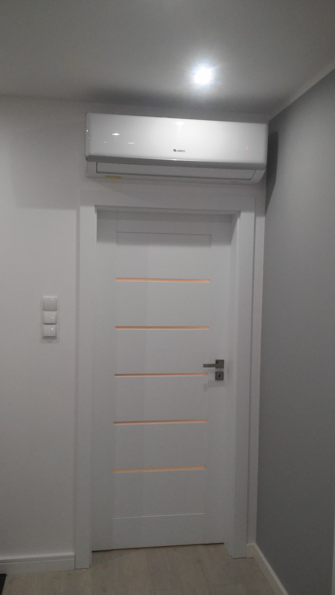 Biała klimatyzacja marki Gree zamontowana nad białą, nowoczesną, wewnętrzną ościeżnicą drzwiową z poziomymi przeszkleniami w jasnym wnętrzu.