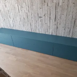 Długa, niska komoda w odcieniach niebieskiego z szufladami, na drewnianej podłodze, na tle ściany z dekoracyjnej cegły.