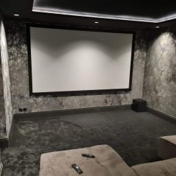 Mateusz Świeczkowski - Nowoczesne kino domowe z dużym ekranem projekcyjnym, ścianami o fakturze betonu, podświetlanym sufitem i wygodną sofą z pilotami.