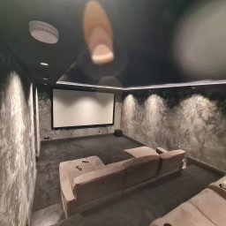 Mateusz Świeczkowski - Wnętrze pokoju kinowego z projektorem, szarą sofą z regulowanymi oparciami i ścianami z efektem betonu, oświetleniem LED i czarnym sufitem.