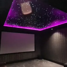Kino domowe z sufitem napinanym imitującym rozgwieżdżone niebo, podświetlonym fioletową listwą LED, oraz ekranem projekcyjnym.