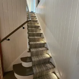 Schody drewniane z wykładziną w szaro-beżową kratę, oświetlone małymi punktowymi lampkami w ścianie, z drewnianą balustradą i boazerią ścienną.