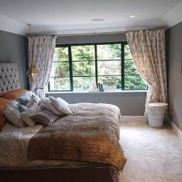 Mateusz Świeczkowski - Sypialnia z szarymi ścianami, dużym oknem z widokiem na zieleń, łóżkiem z pikowanym zagłówkiem i wzorzystymi zasłonami, oświetlona geometrycznymi lampami wiszącymi.