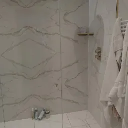 Nowoczesna kabina prysznicowa z deszczownicą, wykończona białymi płytkami imitującymi marmur z szarymi żyłami, szklane drzwi z złotymi zawiasami, mała półka z kosmetykami, ręczniki na grzejniku.