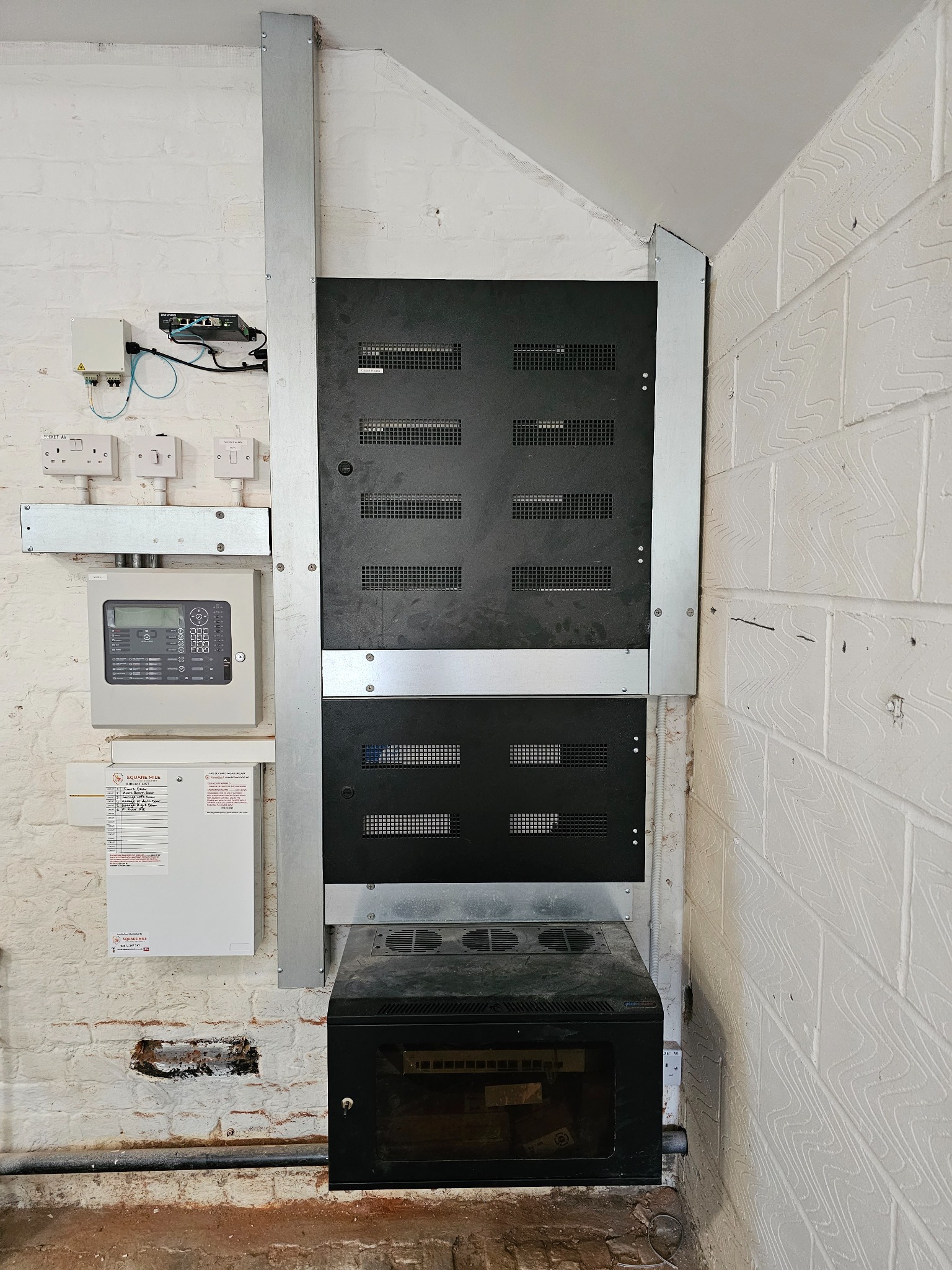 Instalacja szafy serwerowej z metalowymi wspornikami na białej ceglanej ścianie, widoczne okablowanie i panel sterowania alarmem.