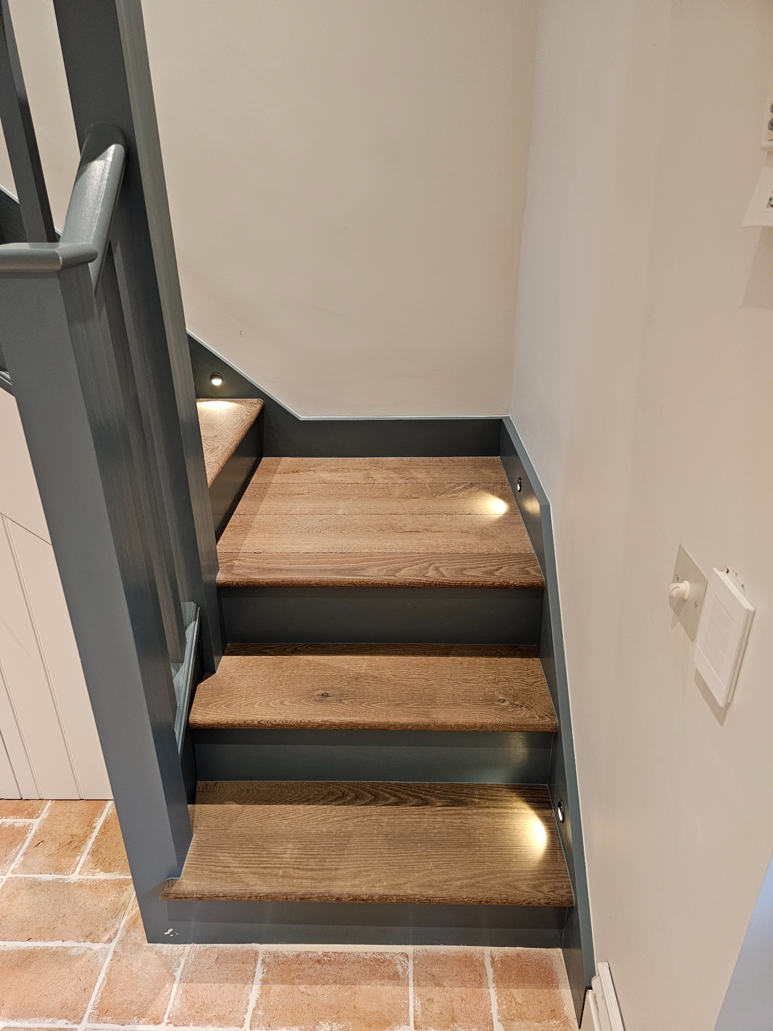 Drewniane schody z podświetleniem LED w ścianie, szara balustrada, podłoga z cegieł, białe ściany.