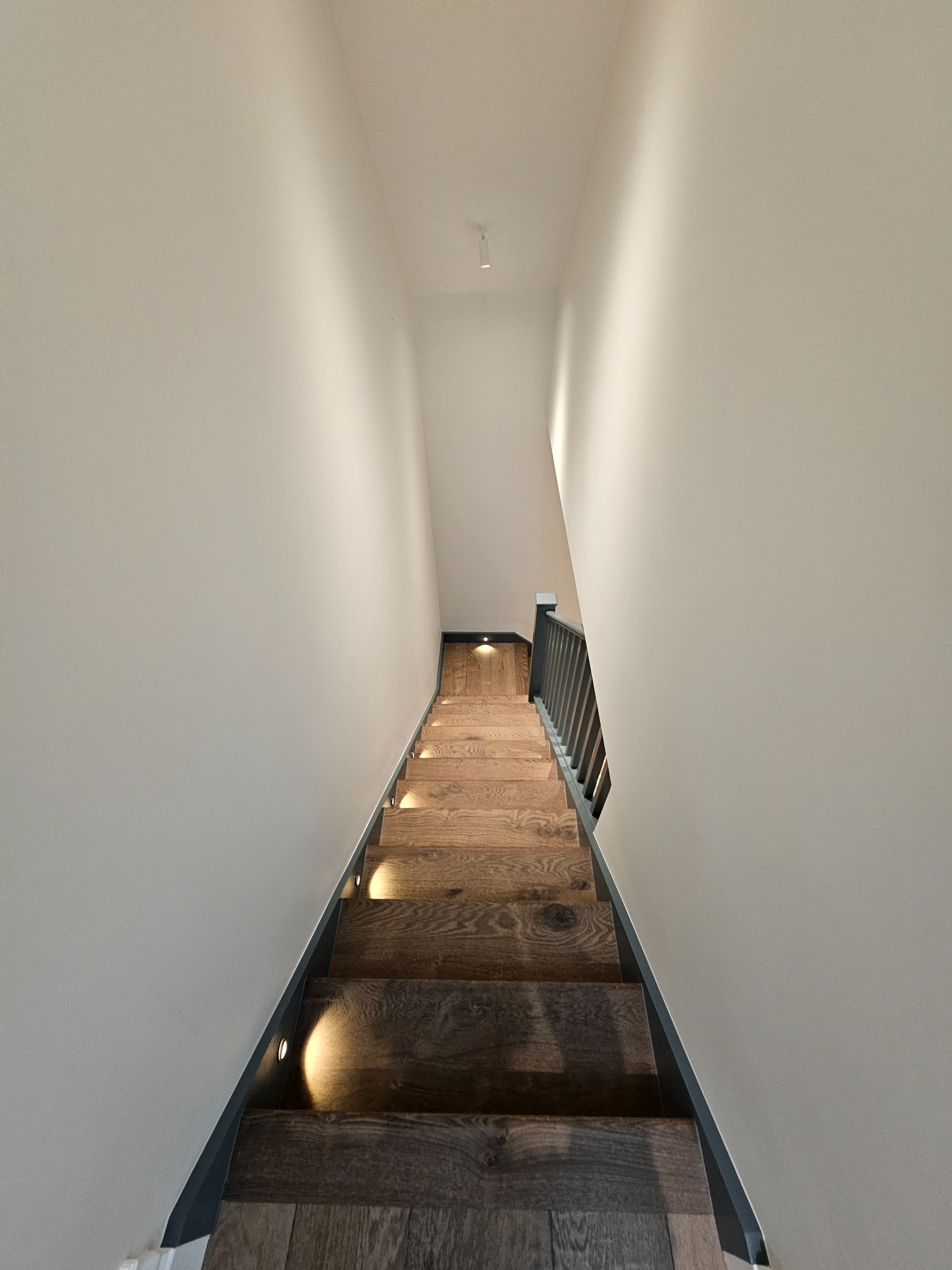 Schody drewniane z oświetleniem LED wbudowanym w stopnie, widok z góry w dół, białe ściany, szara balustrada.