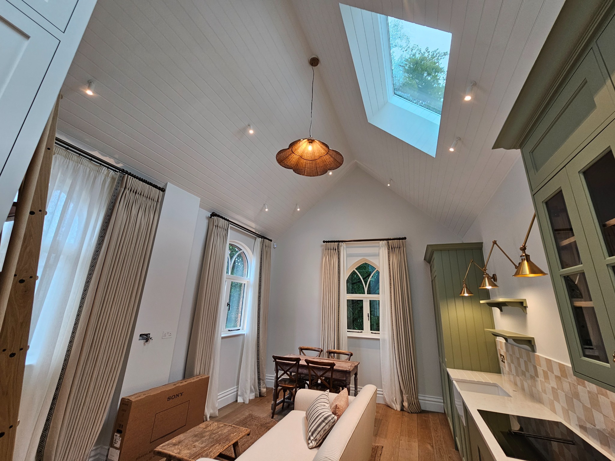 Jasne wnętrze ze skośnym sufitem, oknem dachowym, drewnianą podłogą, zabudową kuchenną w odcieniach zieleni, dekoracyjną lampą i oknami łukowymi.