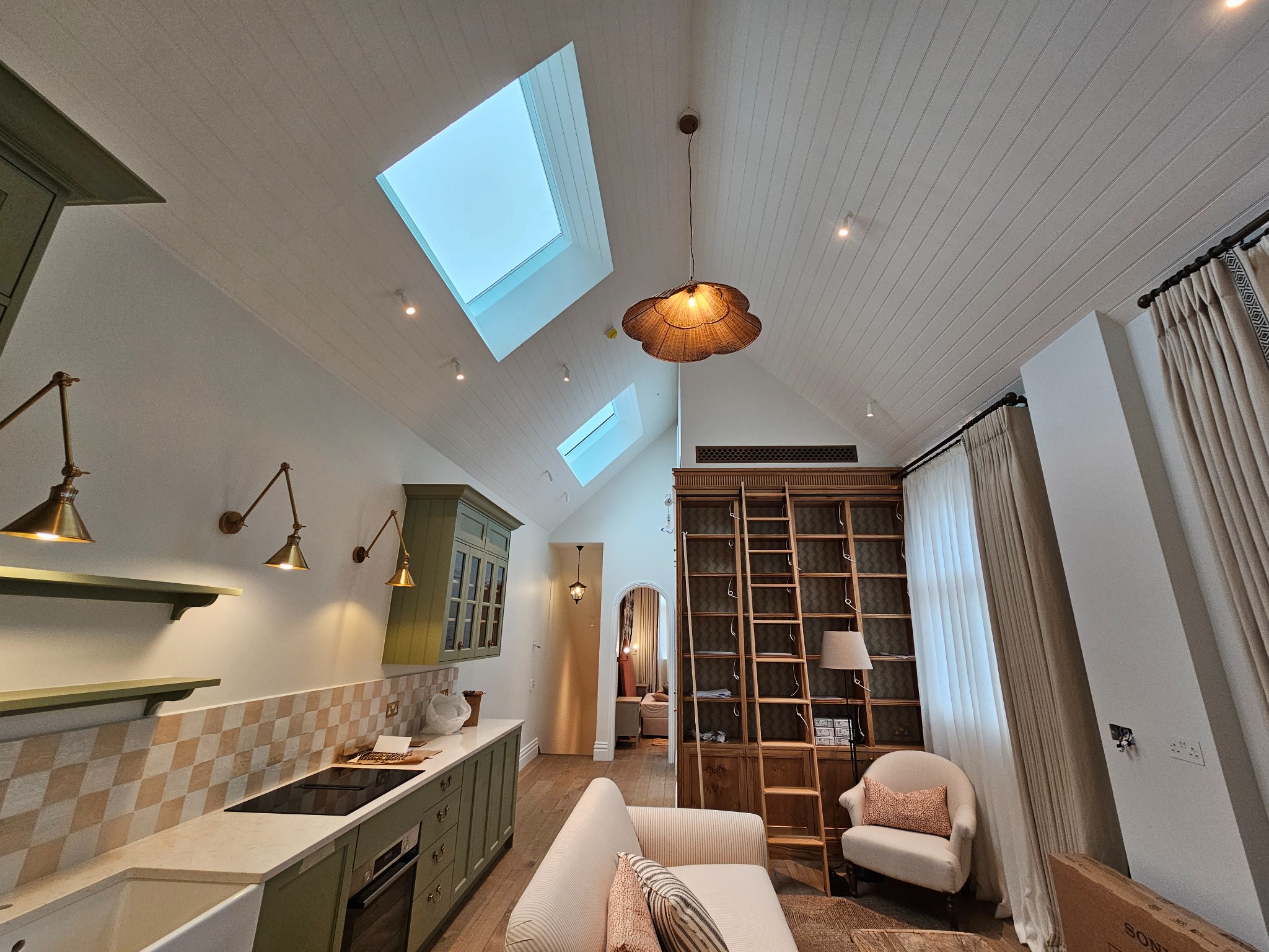 Wnętrze domu z aneksem kuchennym w stylu retro, oświetlone lampami w kolorze starego złota i naturalnym światłem z okien dachowych, z drewnianą biblioteczką z drabiną i beżową sofą w części...