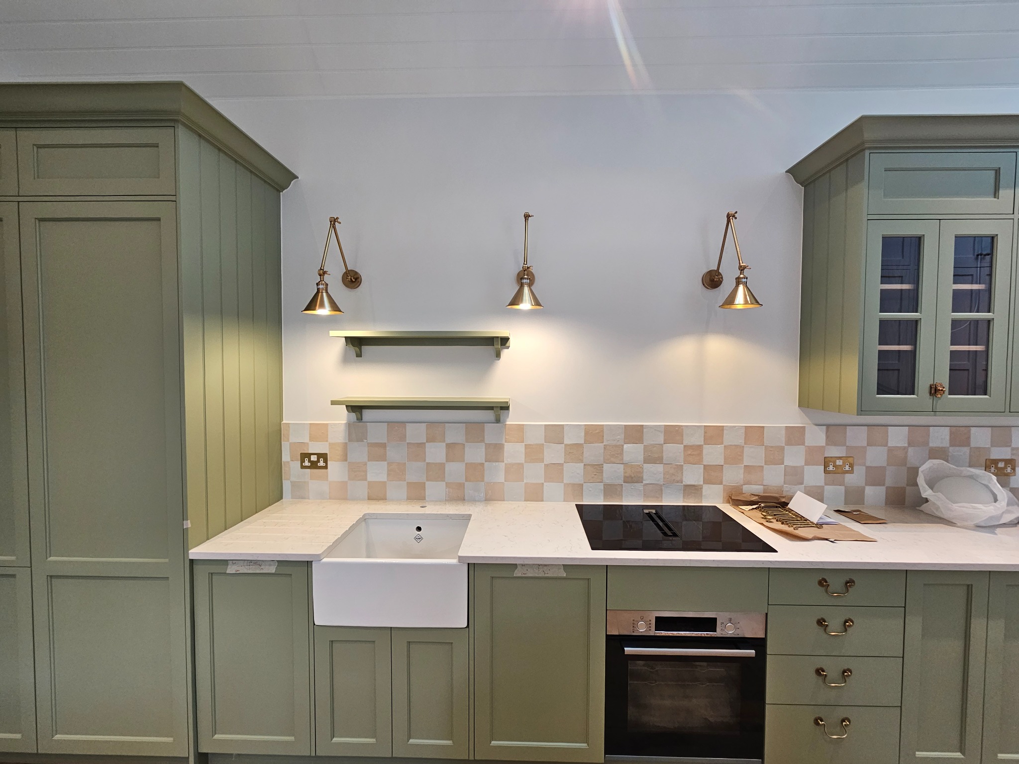 Nowoczesna kuchnia w odcieniach zieleni z białym zlewem farmerskim, płytą indukcyjną i złotymi lampami ściennymi.