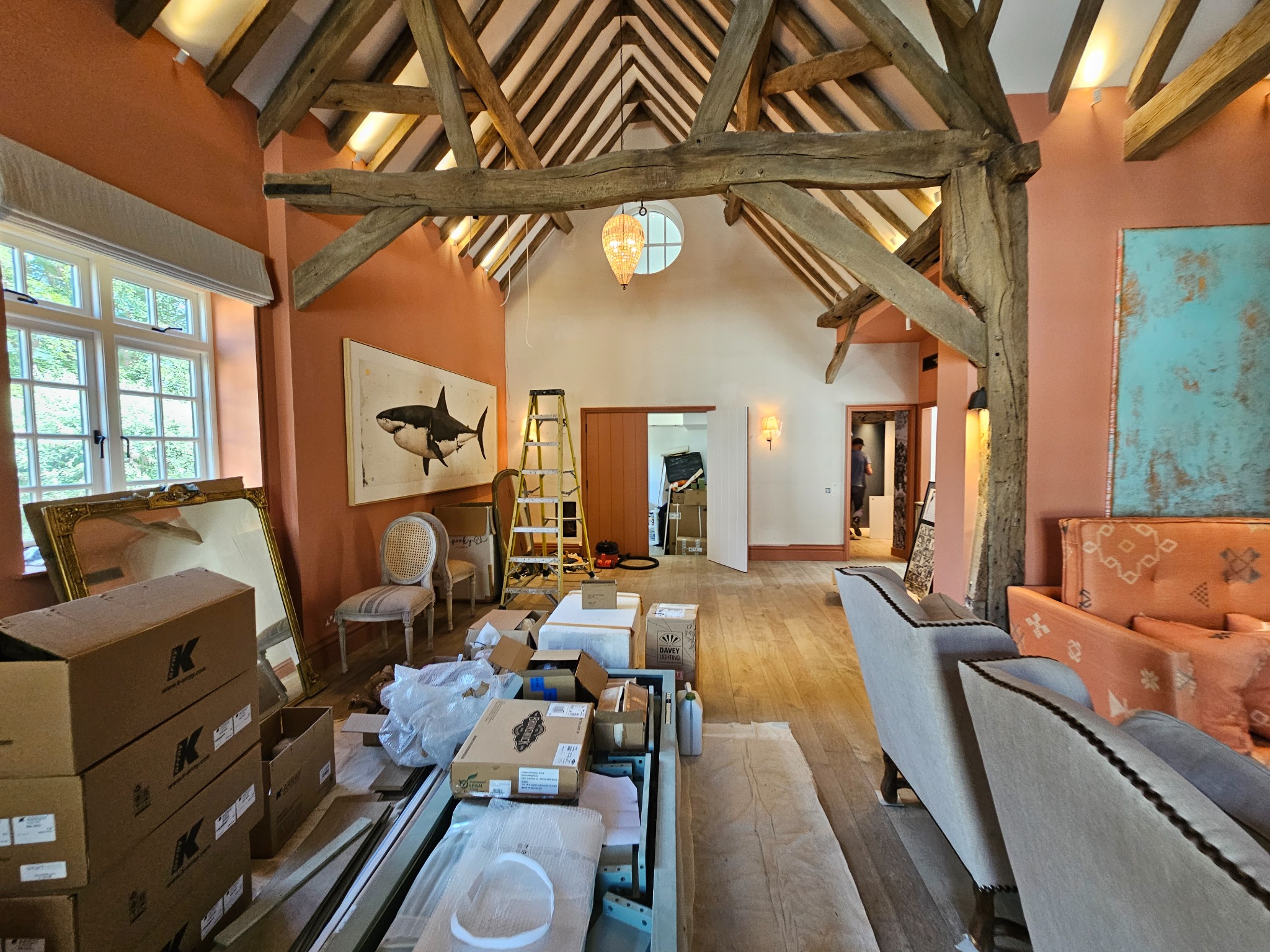 Wnętrze domu z drewnianą konstrukcją stropu, w trakcie prac wykończeniowych, widoczne kartony, meble i drabina, ściany w odcieniach pomarańczy i bieli, okno z widokiem na zieleń.