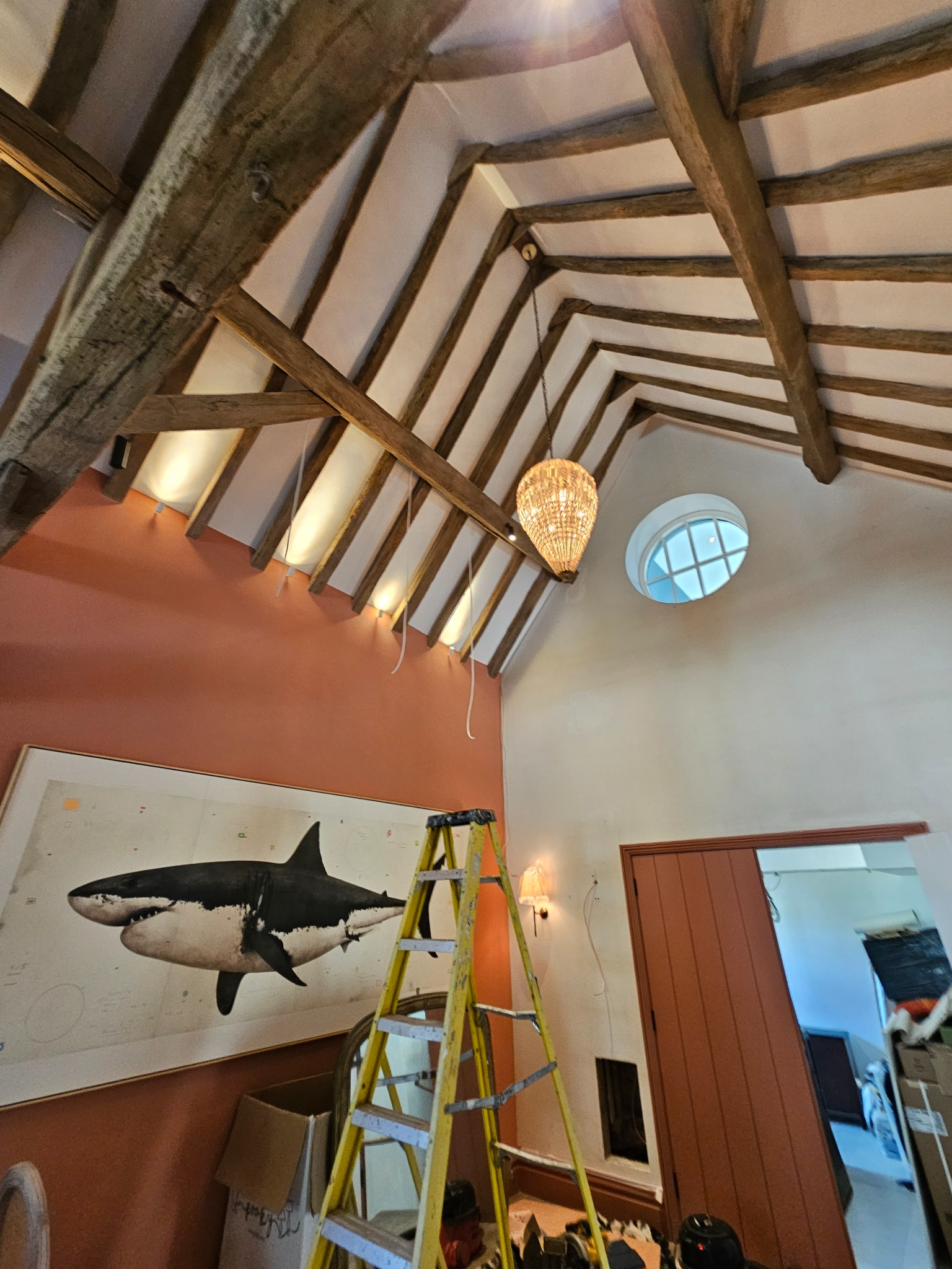Wnętrze pokoju z drewnianą konstrukcją stropu, nową terakotową ścianą, obrazem rekina i wiszącą lampą w kształcie kuli, z żółtą drabiną w tle.