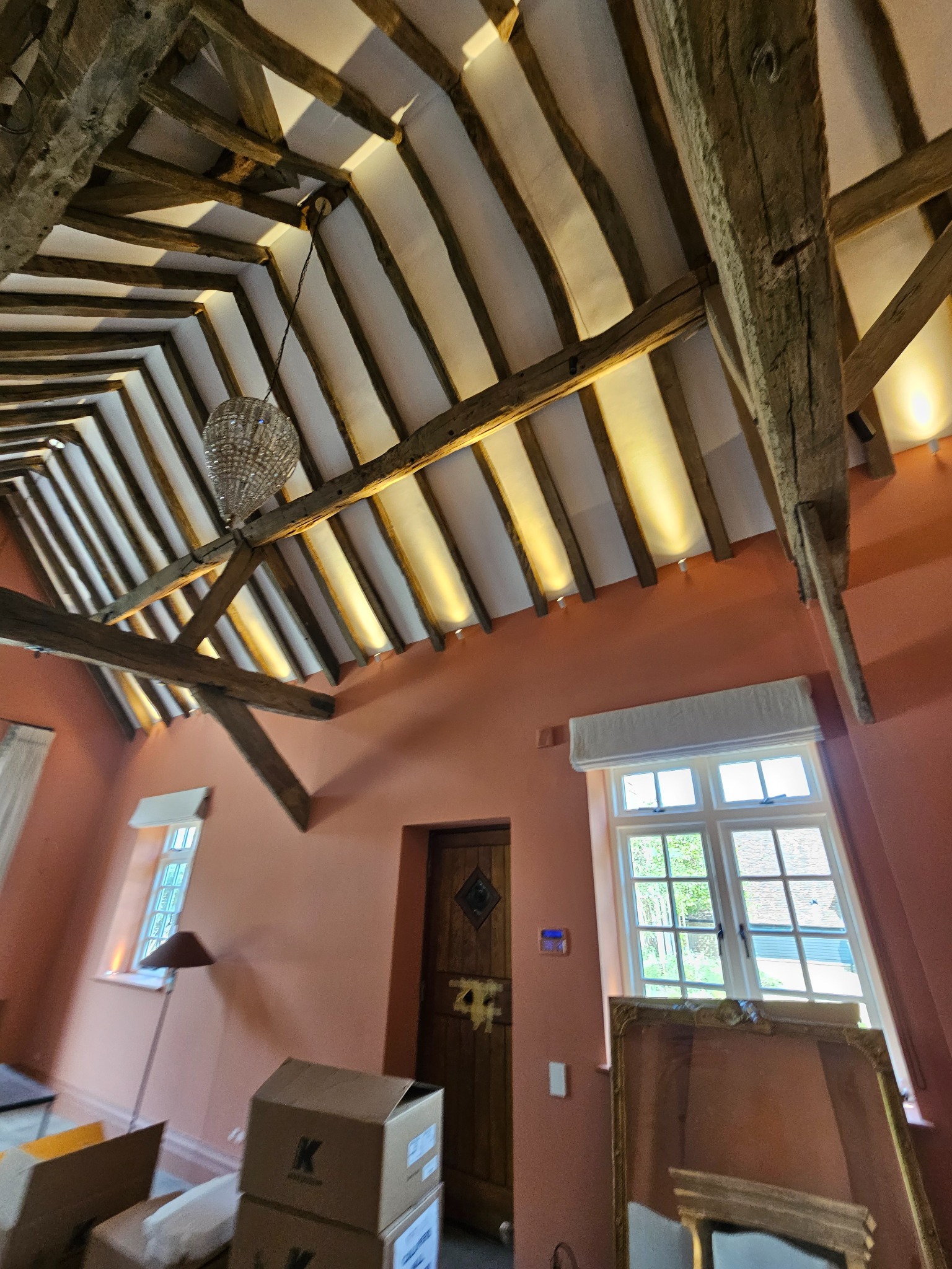 Poddasze z odsłoniętą konstrukcją drewnianą, oświetleniem punktowym i ozdobnym żyrandolem, ściany pomalowane na brzoskwiniowy kolor, widoczne okna i kartony.