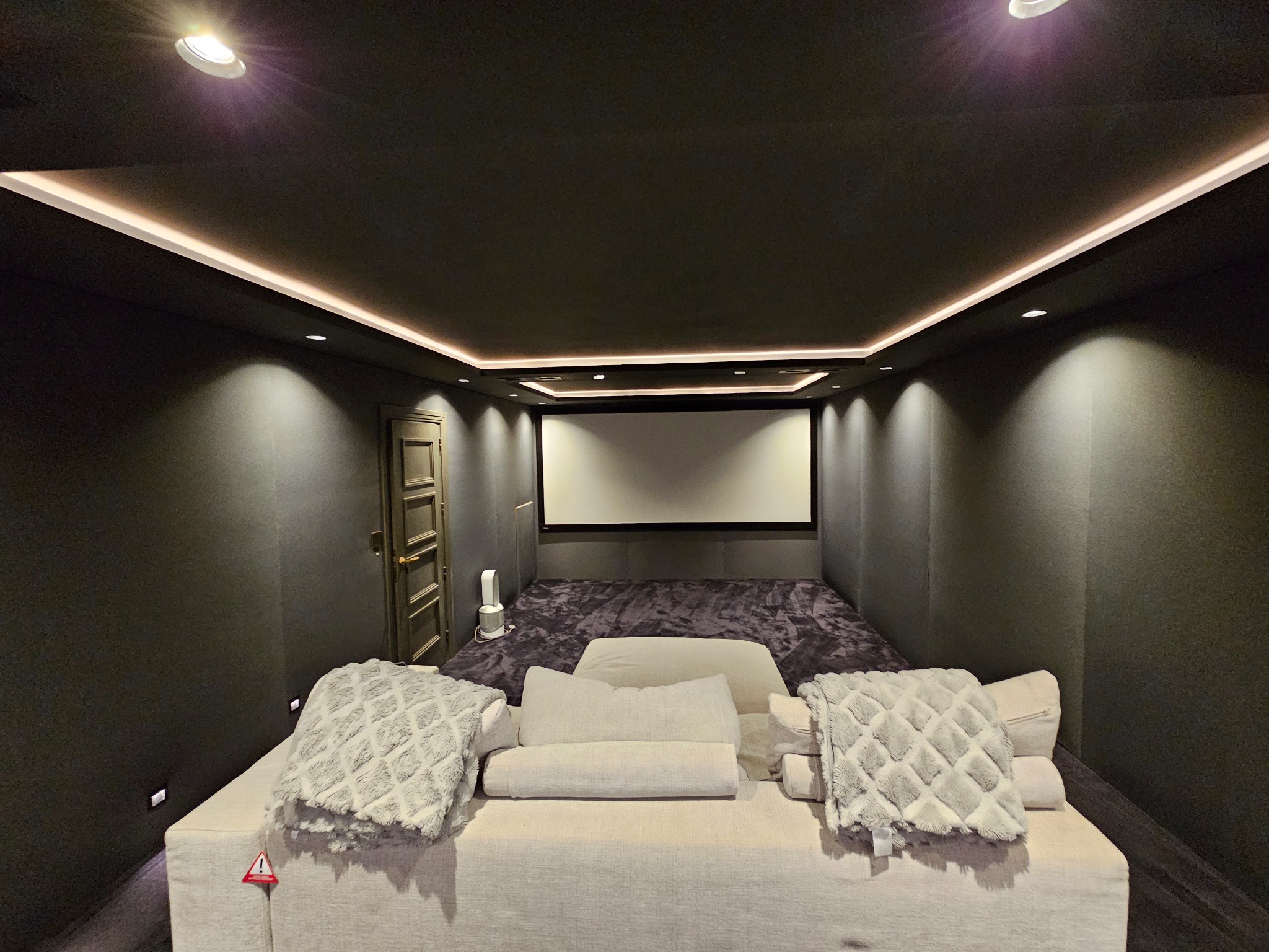 Nowoczesna sala kinowa w domu z ciemnymi ścianami, oświetleniem LED na suficie i dużym ekranem projekcyjnym, widok z wygodnej sofy z poduszkami i pledami.