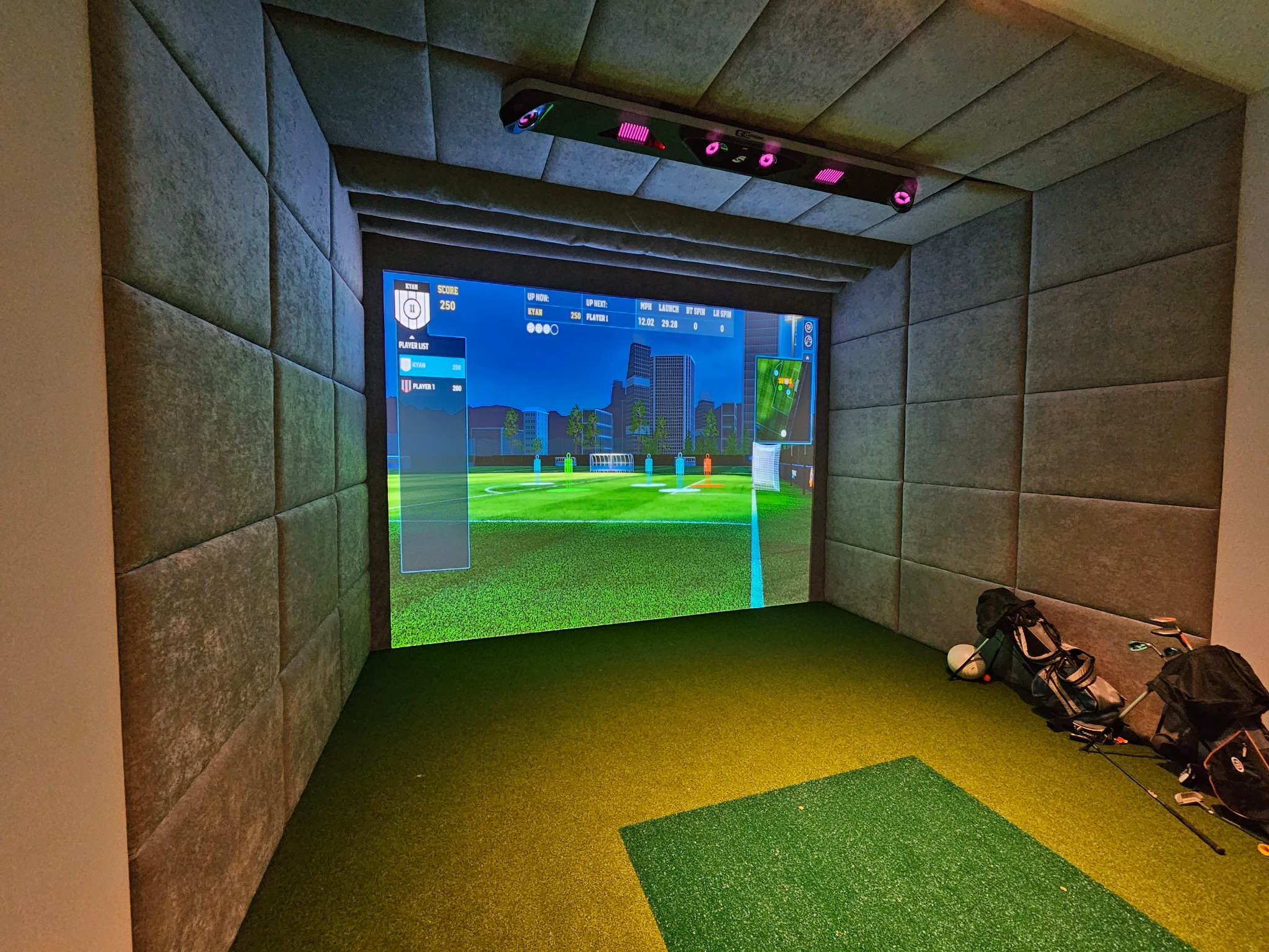Wnętrze pokoju z ekranem symulatora gry w golfa, ściany wyłożone panelami tapicerowanymi, torby golfowe w rogu.