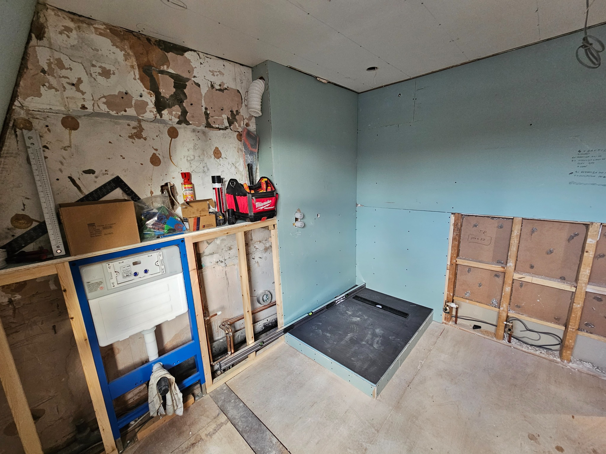 Łazienka w trakcie remontu: zamontowany stelaż podtynkowy WC w niebieskiej ramie, przygotowany brodzik z odpływem liniowym, ściany częściowo wykończone płytami gipsowo-kartonowymi, widoczne...