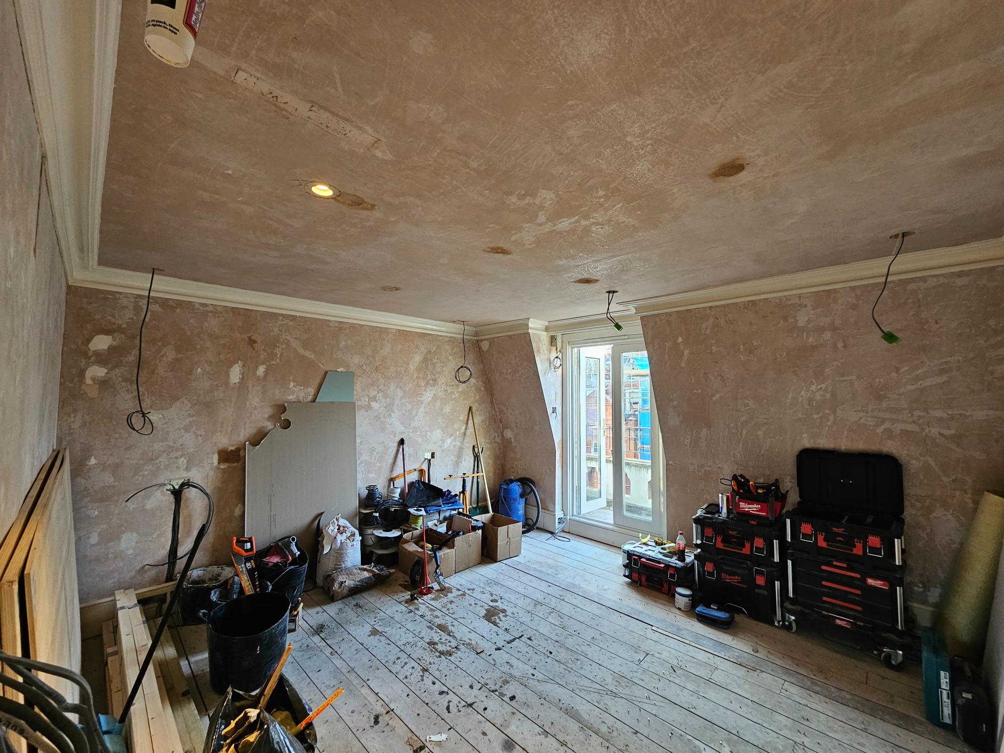 Pokój w trakcie prac wykończeniowych: surowe ściany z widocznymi śladami po tynkowaniu, drewniana podłoga z zabrudzeniami, narzędzia i materiały budowlane rozłożone na podłodze, otwarte okno...