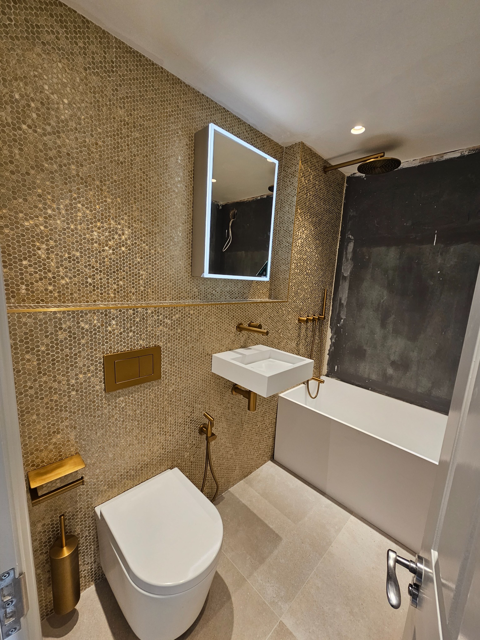 Nowoczesna łazienka w trakcie wykańczania, z mozaiką w odcieniach złota, białą armaturą i złotymi elementami wyposażenia, widoczna surowa ściana obok wanny.